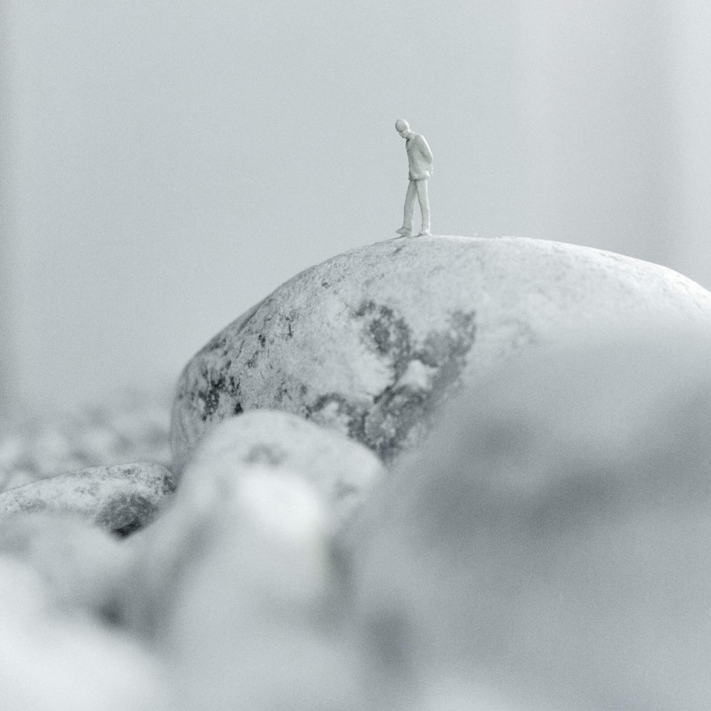 Miniaturfigur eines Menschen auf einem großen Felsen in grauem, minimalistischen Umfeld.